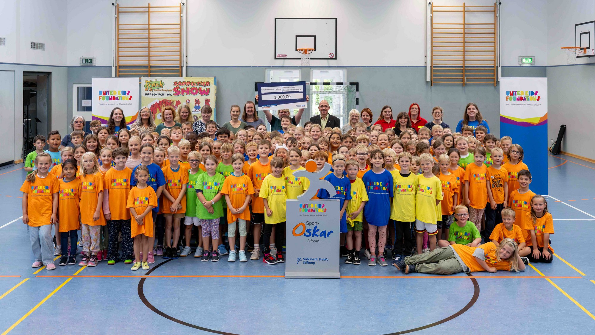 Alle Schülerinnen und Schüler der Grundschule Jembke wurden vom großen Sport-Oskar-Fest motiviert. Credit: Volksbank BRAWO/Isabell Massel Große Gruppe an Kindern und vereinzelt Erwachsene stehen in bunten Tshirts in einer Turnhalle.