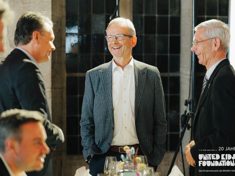 Eine Gruppe von Männern in Anzügen unterhält sich fröhlich bei einer Veranstaltung. Einer von ihnen steht in der Mitte im Fokus, lächelt breit und trägt ein graues Sakko über einem weißen Hemd. Die Atmosphäre wirkt elegant und festlich. Im Hintergrund sind schwarze, quadratische Fenster zu sehen. Unten rechts im Bild befindet sich ein Logo mit der Aufschrift „20 Jahre United Kids Foundations“.