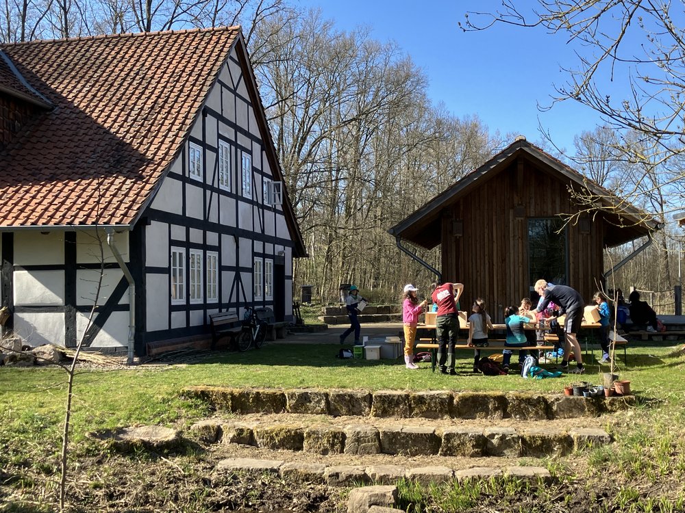 Grünfläche mit Steintreppe, im Hintergrund links ein Fachwerkhaus und rechts eine Holzhütte.