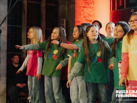 Eine Gruppe von Kindern singt auf einer Bühne. Sie tragen bunte T-Shirts und schauen fröhlich in die gleiche Richtung. Im Hintergrund sind dekorative Wände zu sehen. Links vorne steht ein Mikrofonständer. Die Szene ist in warmem Licht beleuchtet und im Vordergrund befindet sich das Logo "United Kids Foundations", das 20 Jahre feiert.