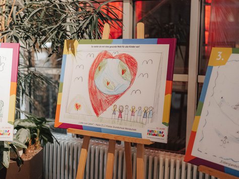 Ein Kinderbild steht auf einer Staffelei in einem Raum mit roten Glasfenstern im Hintergrund. Das Bild zeigt eine Erdkugel, die von einem großen roten Herz umgeben ist. Darunter sind mehrere Personen gemalt. Links im Bild ist eine Pflanze zu sehen, rechts ein weiteres Bild. Auf dem Bild steht: „Liebe zum Leben“ von Maile, 1. Klasse, Grundschule Wendischott. Das Logo der United Kids Foundations ist unten rechts zu sehen.
