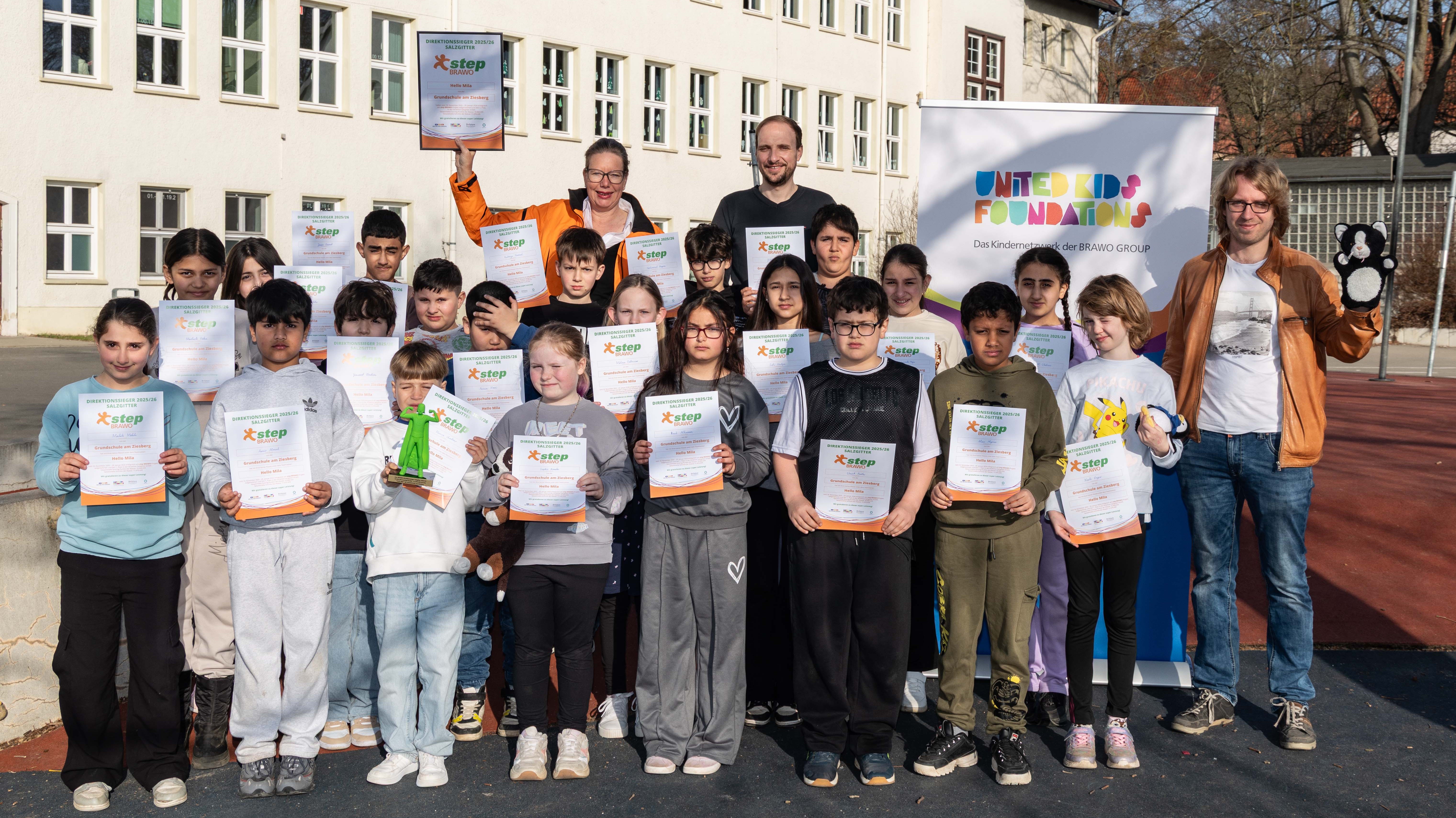 Eine Gruppe von Kindern steht zusammen mit drei Erwachsenen vor einem Schulgebäude. Alle halten Zertifikate mit dem Logo "step" darauf. Hinter ihnen ist ein Banner der "United Kids Foundations" sichtbar. Zwei der Erwachsenen heben ein Plüschtier und ein weiteres Objekt in die Höhe. Die Kinder lächeln in die Kamera, während die Sonne scheint.