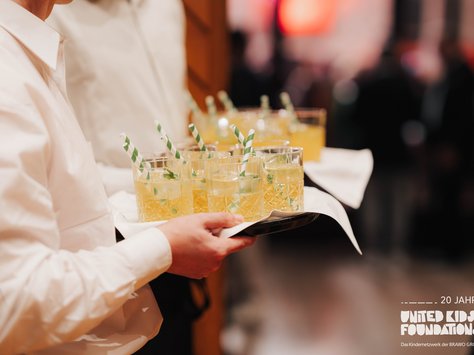 Ein Kellner in weißem Hemd trägt ein Tablett mit mehreren gläsernen Getränken. Die Gläser enthalten ein gelbes Getränk mit grünen Blättern, Eiswürfeln und grün-weiß gestreiften Strohhalmen. Im Hintergrund sind unscharfe Silhouetten von Menschen zu sehen. In der unteren rechten Ecke befindet sich das Logo "United Kids Foundations" mit dem Hinweis auf das 20-jährige Jubiläum der Initiative.