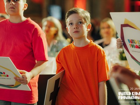 Zwei Kinder stehen nebeneinander in einem Raum mit gedämpftem Licht. Eines trägt ein rotes T-Shirt, das andere ein oranges. Beide halten Plakate mit bunten Logos in den Händen. Im Hintergrund sind unscharf weitere Menschen zu sehen, die an Tischen sitzen. Auf der rechten unteren Ecke des Bildes ist ein Logo mit der Aufschrift „20 Jahre United Kids Foundations“ zu sehen.