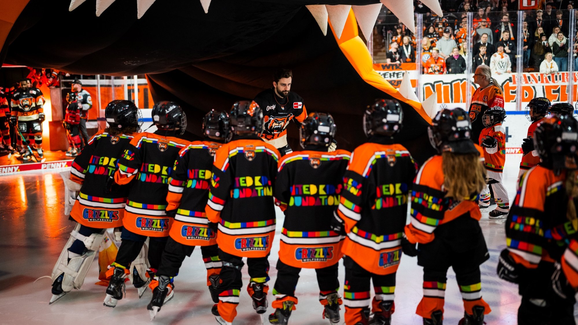 In einer belebten Eishalle steht eine Gruppe junger Eishockeyspieler:innen in bunten Trikots mit der Aufschrift "United Kids Foundation" und "Grizzlys Wolfsburg" auf dem Eis. Sie schauen einem erwachsenen Spieler entgegen, der aus einem großen, aufblasbaren Torbogen mit gezahnten Verzierungen aus dem Mund eines stilisierten Tierkopfes tritt. Im Hintergrund sind weitere Menschen und Spieler zu sehen, während die Zuschauer:innen auf den Tribünen das Geschehen verfolgen.