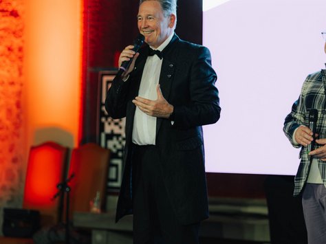 Ein Mann in festlicher Kleidung steht auf einer Bühne und spricht in ein Mikrofon. Der Hintergrund ist warm beleuchtet mit einem orangefarbenen Licht. Rechts im Bild ist ein Ausschnitt einer anderen Person zu sehen. Auf einer Leinwand im Hintergrund steht "United Kids Foundations".