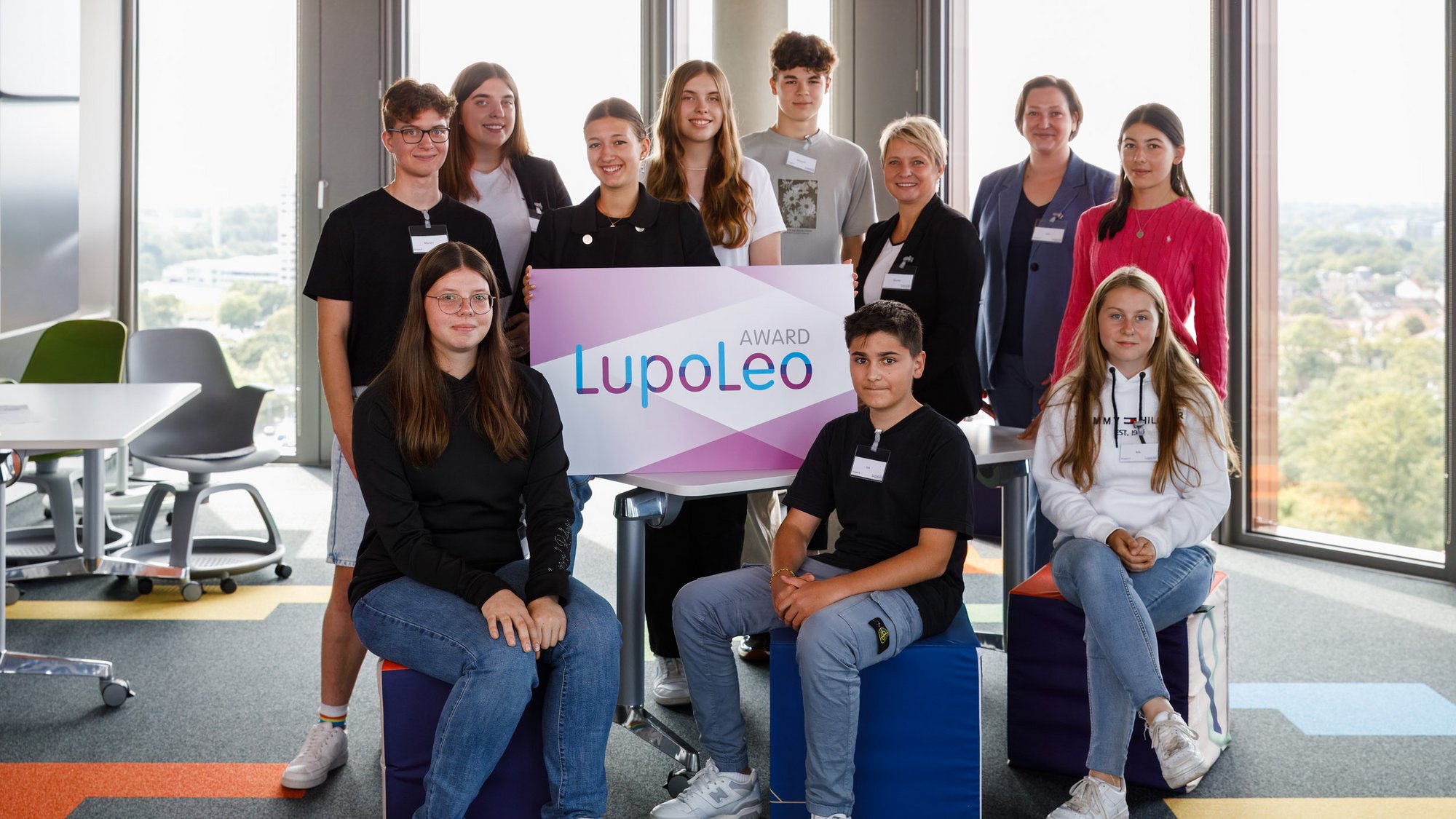 Auf dem Bild ist eine Gruppe junger Menschen abgebildet. Drei der Personen sitzen vor dem Rest der Gruppe auf blauen Hockern. In der Mitte des Bildes hält eine Person ein großes Schild mit der Aufschrift "Award - LupoLeo". Die Gruppe steht in einem Büro, im Hintergrund sind große Fensterscheiben.