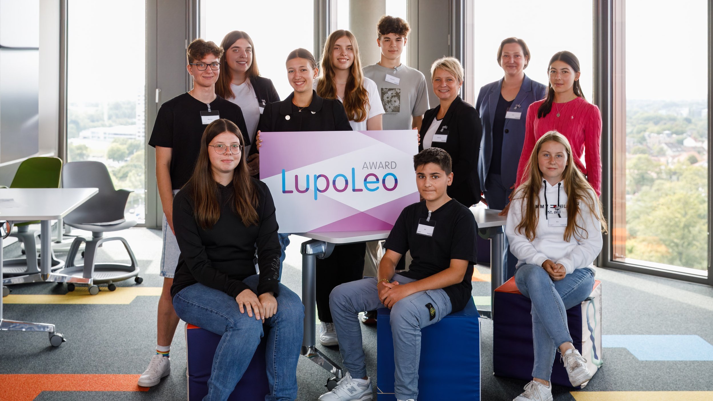 Auf dem Bild ist eine Gruppe junger Menschen abgebildet. Drei der Personen sitzen vor dem Rest der Gruppe auf blauen Hockern. In der Mitte des Bildes hält eine Person ein großes Schild mit der Aufschrift "Award - LupoLeo". Die Gruppe steht in einem Büro, im Hintergrund sind große Fensterscheiben.