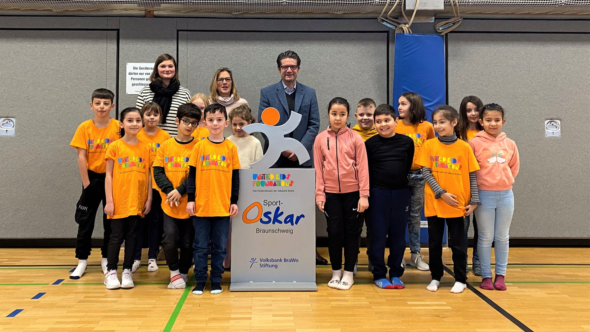 Gruppe von Kindern in orangenen Shirts mit drei Erwachsenen stehen lächelnd in einer Sporthalle.