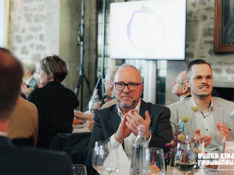 Eine Gruppe von Menschen sitzt an einem Tisch in einem Veranstaltungsraum. Im Vordergrund klatschen zwei Männer, einer trägt eine Brille. Auf dem Tisch stehen Wasserflaschen und Weingläser. Im Hintergrund ist ein verschwommener Bildschirm mit einem Jubiläumslogo zu sehen. An der Wand hängt ein großes Bild. Der Text am unteren rechten Bildrand weist auf das 20-jährige Jubiläum der United Kids Foundations hin.