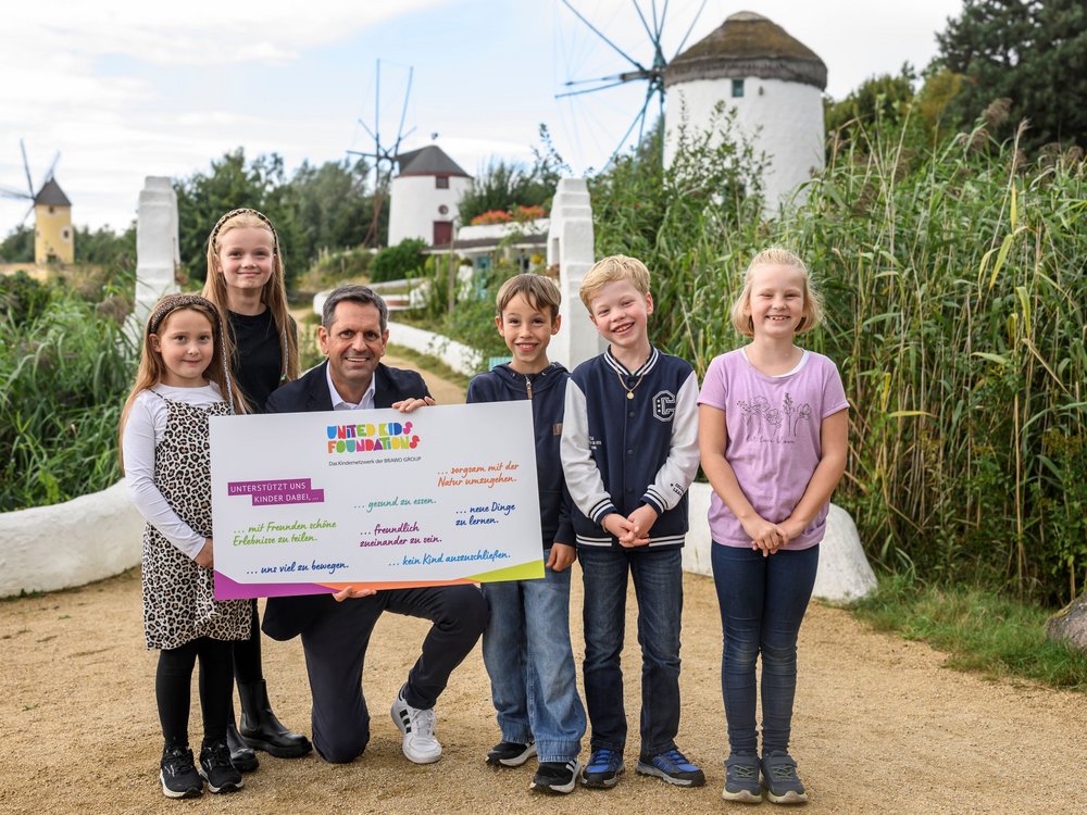 Eine Gruppe von fünf Kindern und einem Erwachsenen posiert draußen vor einer Kulisse mit Windmühlen und grüner Vegetation. Der Erwachsene hält ein buntes Plakat mit der Aufschrift "United Kids Foundations" und verschiedenen unterstützenden Botschaften für Kindererfahrungen. Die Stimmung ist fröhlich und die Natur um sie herum wirkt friedlich.