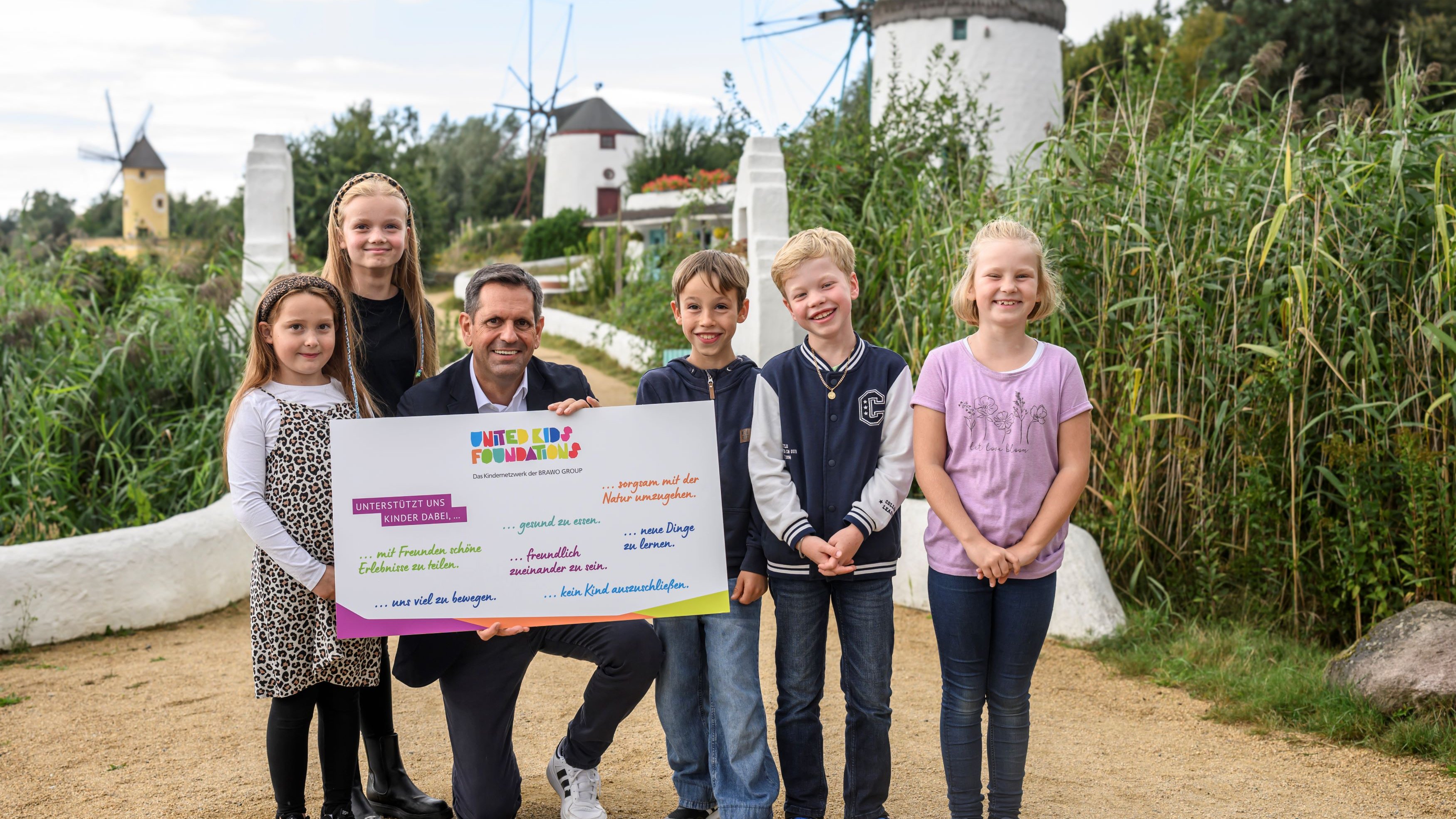 Eine Gruppe von fünf Kindern und einem Erwachsenen posiert draußen vor einer Kulisse mit Windmühlen und grüner Vegetation. Der Erwachsene hält ein buntes Plakat mit der Aufschrift "United Kids Foundations" und verschiedenen unterstützenden Botschaften für Kindererfahrungen. Die Stimmung ist fröhlich und die Natur um sie herum wirkt friedlich.