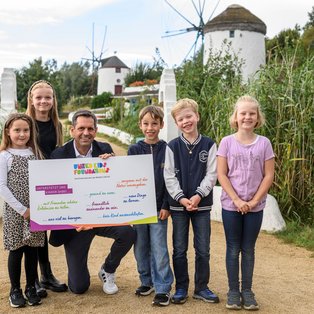 Eine Gruppe von fünf Kindern und einem Erwachsenen posiert draußen vor einer Kulisse mit Windmühlen und grüner Vegetation. Der Erwachsene hält ein buntes Plakat mit der Aufschrift "United Kids Foundations" und verschiedenen unterstützenden Botschaften für Kindererfahrungen. Die Stimmung ist fröhlich und die Natur um sie herum wirkt friedlich.