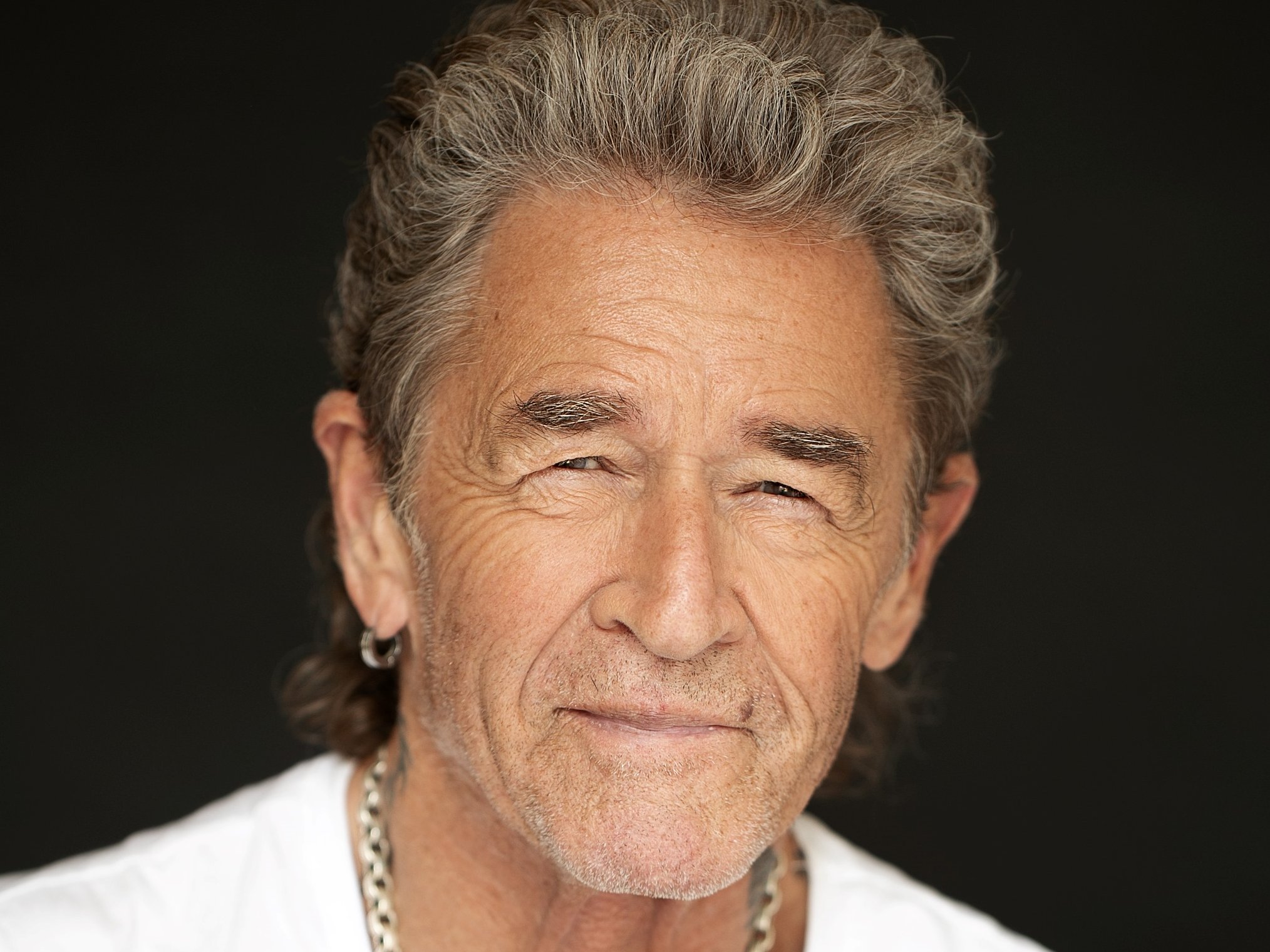 Ein älterer Mann mit grauen Haaren und Bart trägt ein weißes T-Shirt und eine silberne Kette. Er lächelt leicht und hat ein Tattoo auf dem Arm. Der Hintergrund ist dunkel.