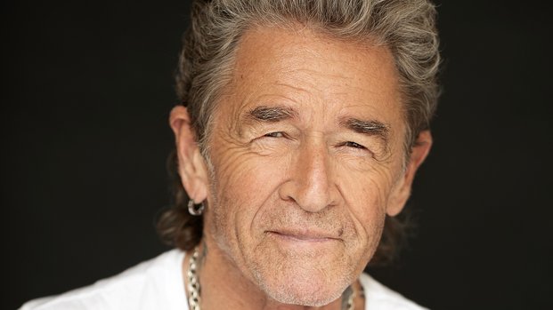 Ein älterer Mann mit grauen Haaren und Bart trägt ein weißes T-Shirt und eine silberne Kette. Er lächelt leicht und hat ein Tattoo auf dem Arm. Der Hintergrund ist dunkel.