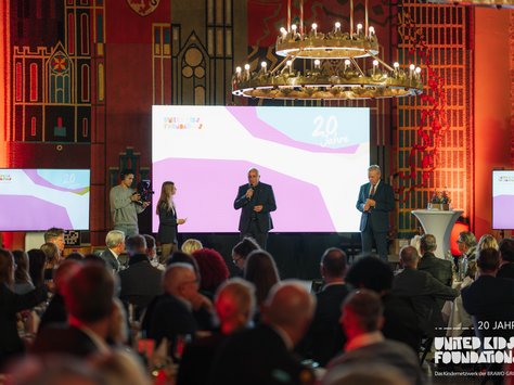 Das Bild zeigt eine Veranstaltung in einem festlich beleuchteten Raum. Im Vordergrund sitzt ein Publikum an Tischen. Auf der Bühne stehen zwei Männer in Anzügen, die in Mikrofone sprechen, während eine Frau daneben steht. Im Hintergrund ist ein großer Monitor zu sehen, der "20 Jahre" und "United Kids Foundations" anzeigt. Links im Bild steht ein Kameramann, der die Szene filmt. Die Kulisse ist bunt mit einem großen Kronleuchter von der Decke und dekorativen Wandmustern.