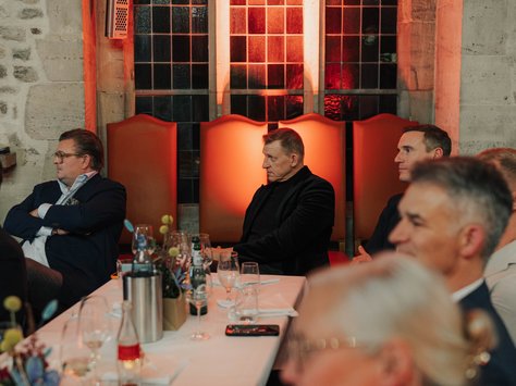 In einem gedämpft beleuchteten Raum sitzen mehrere Personen an einem festlich gedeckten Tisch. Im Hintergrund ist eine große gotische Fensterfront mit warmem orangefarbenem Licht zu sehen. Auf dem Tisch stehen Getränke und Dekorationen. Am unteren Bildrand ist ein Logo mit „20 Jahre United Kids Foundations“ zu erkennen, begleitet von dem Text „Das Kinderetzwerk der Brawo Group“. Die Anwesenden scheinen aufmerksam einer Veranstaltung oder Rede zu folgen.