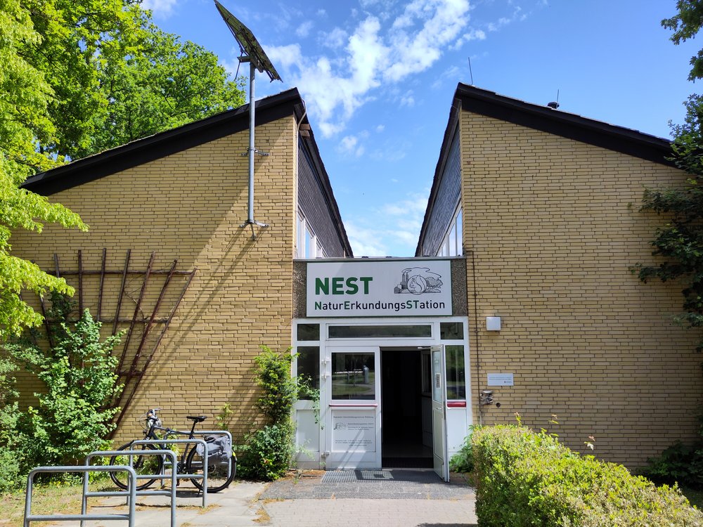 Das Bild zeigt ein Gebäude. Mittig ist der Eingang, über dem ein Schild mit der Aufschrift "NEST NaturErkundungsStation" hängt.