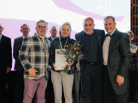 Eine Gruppe von acht Personen steht auf einer Bühne vor einem Hintergrund mit der Aufschrift "20 Jahre United Kids Foundations". Die vier Personen vorne halten eine Urkunde und einen Blumenstrauß. Alle tragen formelle Kleidung. Im Hintergrund sind elegante Wandmuster zu sehen. In der Ecke des Bildes befinden sich Schriftzüge, die für das 20-jährige Jubiläum der United Kids Foundations stehen, ein Kinderhilfsnetzwerk der BraWo Group.