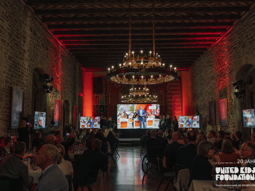 Eine festliche Veranstaltung in einem historischen Saal mit Steinwänden und Holzdecke. Der Raum ist in warmem Rotlicht erleuchtet. Über der Bühne hängen große, runde Kronleuchter. Auf der Bühne spricht eine Person vor einem Bildschirm mit verschiedenen Bildern. Die Gäste sitzen an runden Tischen, einige fotografieren das Geschehen. Im Vordergrund ist das Logo der “United Kids Foundations” zu sehen.