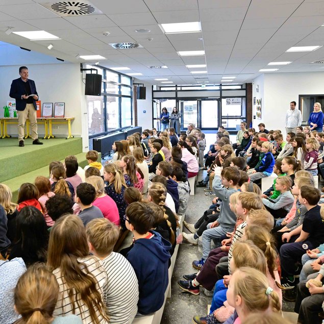 Ein Mann in einem Anzug und ein weiterer Mann stehen auf einer kleinen Bühne und sprechen zu einer großen Gruppe von sitzenden Kindern. Die Kinder sind aufmerksam und schauen zur Bühne. Im Hintergrund sind Banner und Plakate mit Logos zu sehen. Der Raum ist hell erleuchtet und hat große Fenster, die viel Tageslicht hereinlassen. Einige Erwachsene stehen an der Seite und beobachten die Szenerie.