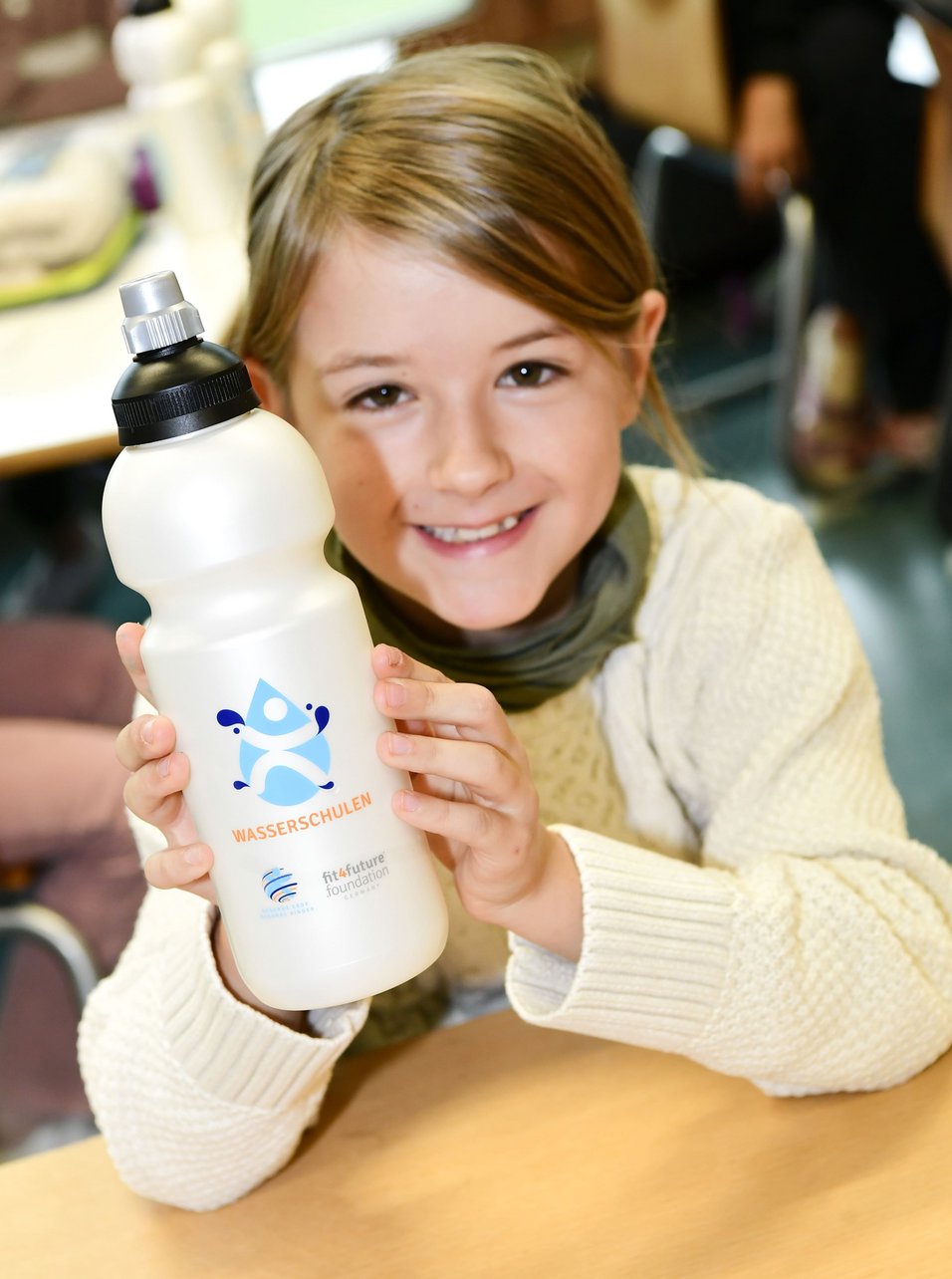 Ein Kind hält lächelnd eine wiederverwendbare Wasserflasche in die Kamera. Auf der Flasche ist ein blaues Logo mit der Aufschrift "Wasserschulen". Unten stehen die Begriffe "fit4future foundation" und "Wasserschulen" zusammen mit weiteren Logos. Im Hintergrund sind unscharf weitere Flaschen und eine Tischfläche zu sehen.