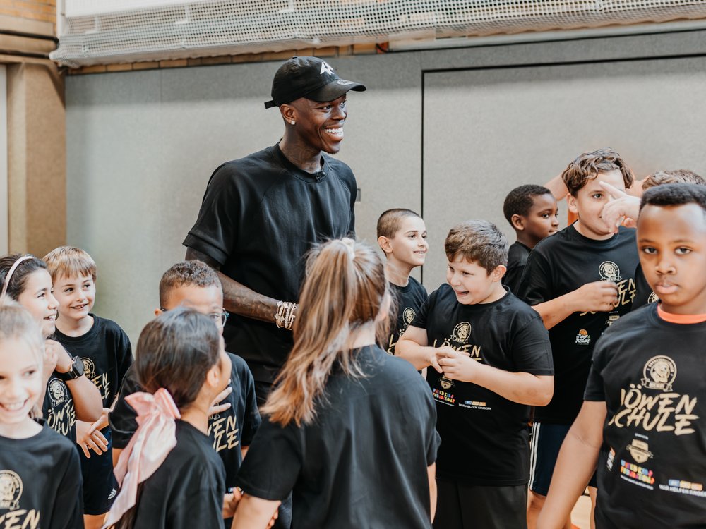 Ein lächelnder Mann in schwarzem Outfit steht inmitten einer fröhlichen Gruppe von Kindern in einer Halle.