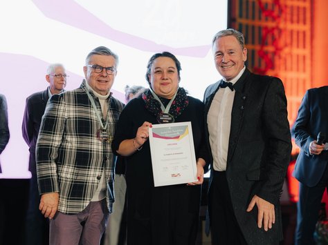 Eine Gruppe von drei Personen steht zusammen und lächelt in die Kamera. Eine Frau in der Mitte hält ein Zertifikat in den Händen. Die Personen tragen formelle Kleidung. Im Hintergrund ist eine Bühne mit einem großen Display zu sehen, auf dem "20 Jahre" steht. Rechts unten im Bild ist das Logo von "United Kids Foundations" zu erkennen.