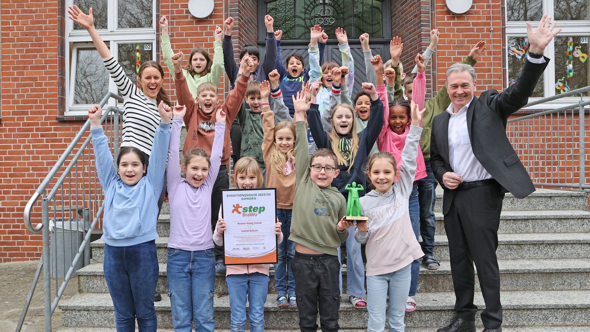 Eine Gruppe Kinder stehen mit zwei Erwachsenen auf einer Treppe und strecken lachend ihre Arme in die Luft. Ein Kind in der ersten Reihe hält eine gerahmte Auszeichnung in der Hand.
