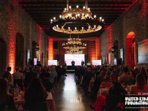 Ein großer, festlich beleuchteter Saal mit hohen Decken und steinernen Wänden. Der Raum ist gefüllt mit vielen Menschen, die an Tischen sitzen, und nach vorne auf eine Bühne schauen. Auf der Bühne stehen drei Personen vor einem großen Bildschirm. Der Saal ist stimmungsvoll in warmen Rot- und Orangetönen beleuchtet, und von der Decke hängen große, verzierte Kronleuchter. Im Vordergrund sieht man Tische mit Flaschen und Gläsern. Links und rechts an den Wänden sind mehrere flache Bildschirme angebracht.