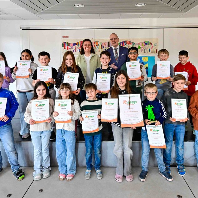 Eine Gruppe von Schülerinnen und Schülern steht auf einer Bühne und hält Urkunden in den Händen. Im Hintergrund sind zwei Erwachsene zu sehen. Die Kinder wirken fröhlich und stolz. Im Raum sind weiße Wände und ein kleines Kunstwerk an der Wand sichtbar. Einige tragen bunte Kleidung, die Stimmung ist positiv.