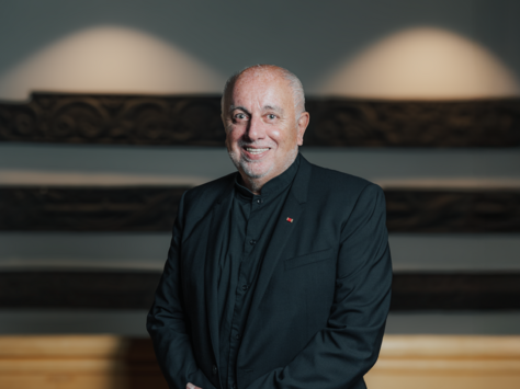 Ein älterer Mann in einem schwarzen Anzug lächelt freundlich in die Kamera. Der Hintergrund ist leicht unscharf und zeigt eine kunstvoll gestaltete Wand. In der unteren rechten Ecke des Bildes befindet sich das Logo von United Kids Foundations mit dem Hinweis auf das 20-jährige Jubiläum.