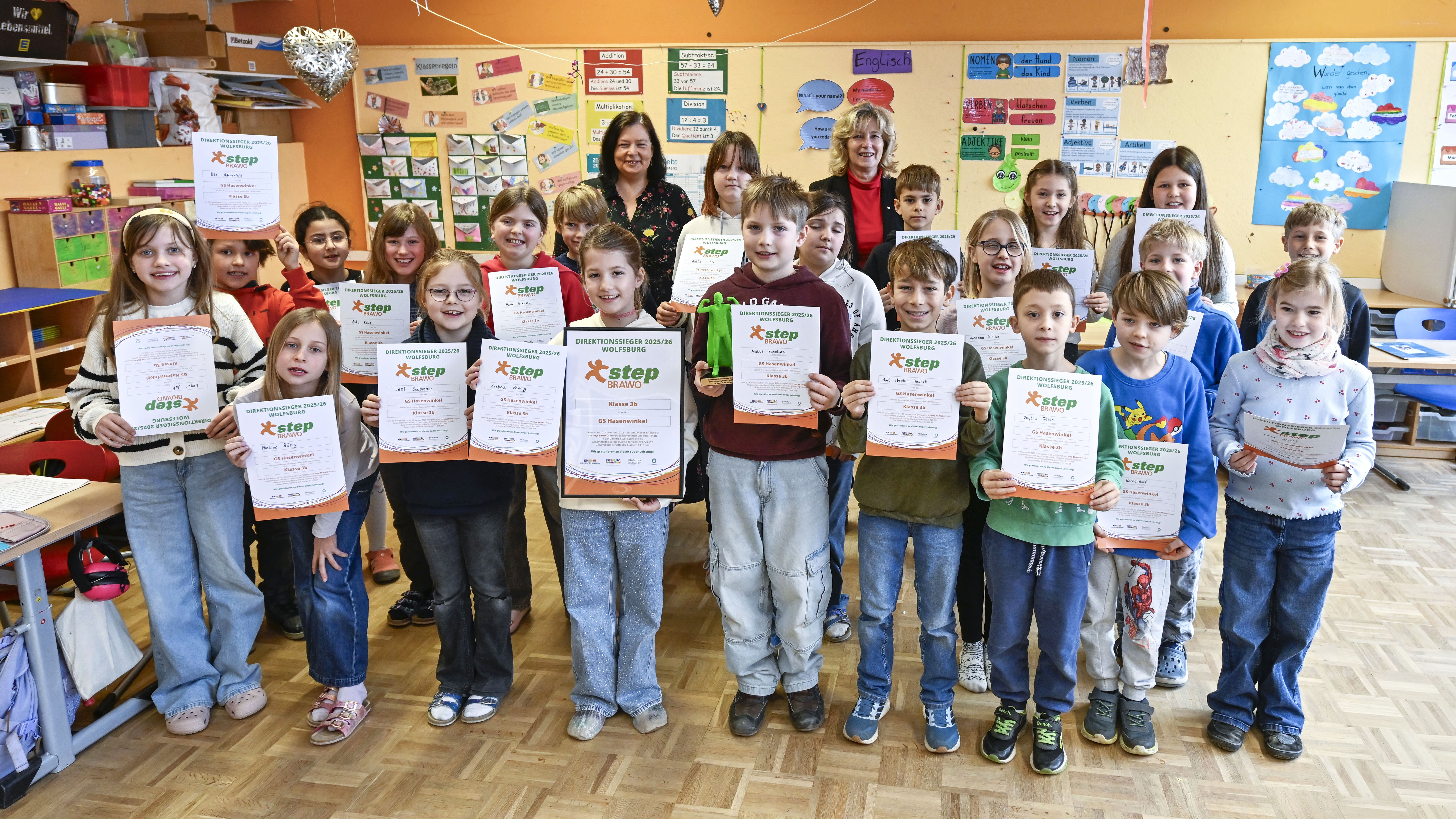 Eine Gruppe von Kindern steht in einem Klassenzimmer und hält stolz Zertifikate in den Händen. Im Hintergrund sind zwei Erwachsene zu sehen. An den Wänden hängen bunte Poster und Schilder. Die Kinder lächeln in die Kamera, während sie ihre Auszeichnungen präsentieren. Der Raum ist bunt dekoriert und es herrscht eine freudige Atmosphäre.