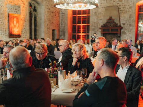 In einem festlich beleuchteten Raum mit alten Steinmauern und Holzbalkendecke sitzen mehrere elegant gekleidete Personen an runden Tischen. Die Tische sind mit weißen Tischdecken, Kerzen, Blumenarrangements und Gläsern dekoriert. Im Hintergrund befindet sich ein großer hängender Kronleuchter. An einer Wand ist ein Kamin zu erkennen. Die Stimmung wirkt aufmerksam und konzentriert. In der unteren Ecke des Bildes ist das Logo der „United Kids Foundations“ zu sehen, begleitet von einem Hinweis auf das 20-jährige Jubiläum des Netzwerks.