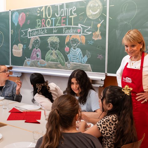 Eine Gruppe von Kindern sitzt an einem Tisch und isst. Im Hintergrund ist eine Tafel zu sehen, auf die mit bunter Kreide gezeichnete Figuren und der Schriftzug „10 Jahre Brotzeit – Frühstück für Kinder“ geschrieben sind. Eine Frau steht daneben und trägt eine rote Schürze mit dem Logo von "brotZeit". Sie lächelt und schaut zu den Kindern. Die Stimmung wirkt fröhlich und einladend.