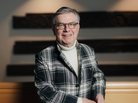 Ein älterer Mann steht lächelnd vor einer Wand. Er trägt ein kariertes Sakko, einen hellen Rollkragenpullover und eine Brille. Im Hintergrund sind Holzelemente an der Wand angebracht. Unten rechts im Bild befindet sich ein Text: "20 Jahre United Kids Foundations, das Kindernetzwerk der Brawo Group."