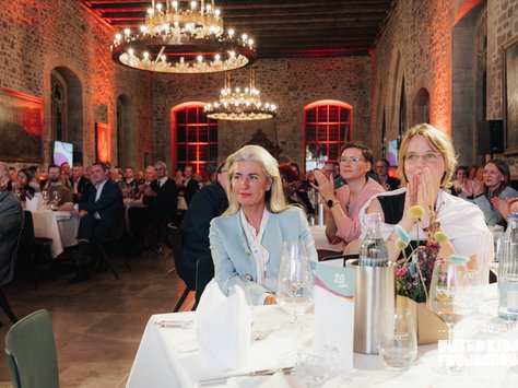 In einem festlich dekorierten Saal mit Steinwänden und großen Kronleuchtern sitzen zahlreiche Menschen an runden Tischen. Eine Frau in einem hellblauen Blazer steht im Vordergrund, umgeben von anderen Gästen, die applaudieren. Die Tische sind mit Weinflaschen, Gläsern und Blumendekorationen geschmückt. Es scheint ein besonderes Event oder eine Feier stattzufinden.
