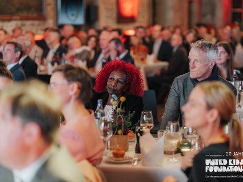 Eine Gruppe von Menschen sitzt an dekorierten Tischen in einem Veranstaltungsraum. In der Mitte sitzt eine Person mit auffälligem roten Haar, aufmerksam in Richtung Bühne blickend. Auf den Tischen stehen Wein- und Wassergläser sowie Blumendekorationen. Im Hintergrund sind weitere Personen zu sehen, die ebenfalls nach vorn schauen. Auf dem Bild ist auch das Logo "United Kids Foundations" und ein Hinweis auf das 20-jährige Jubiläum zu erkennen.
