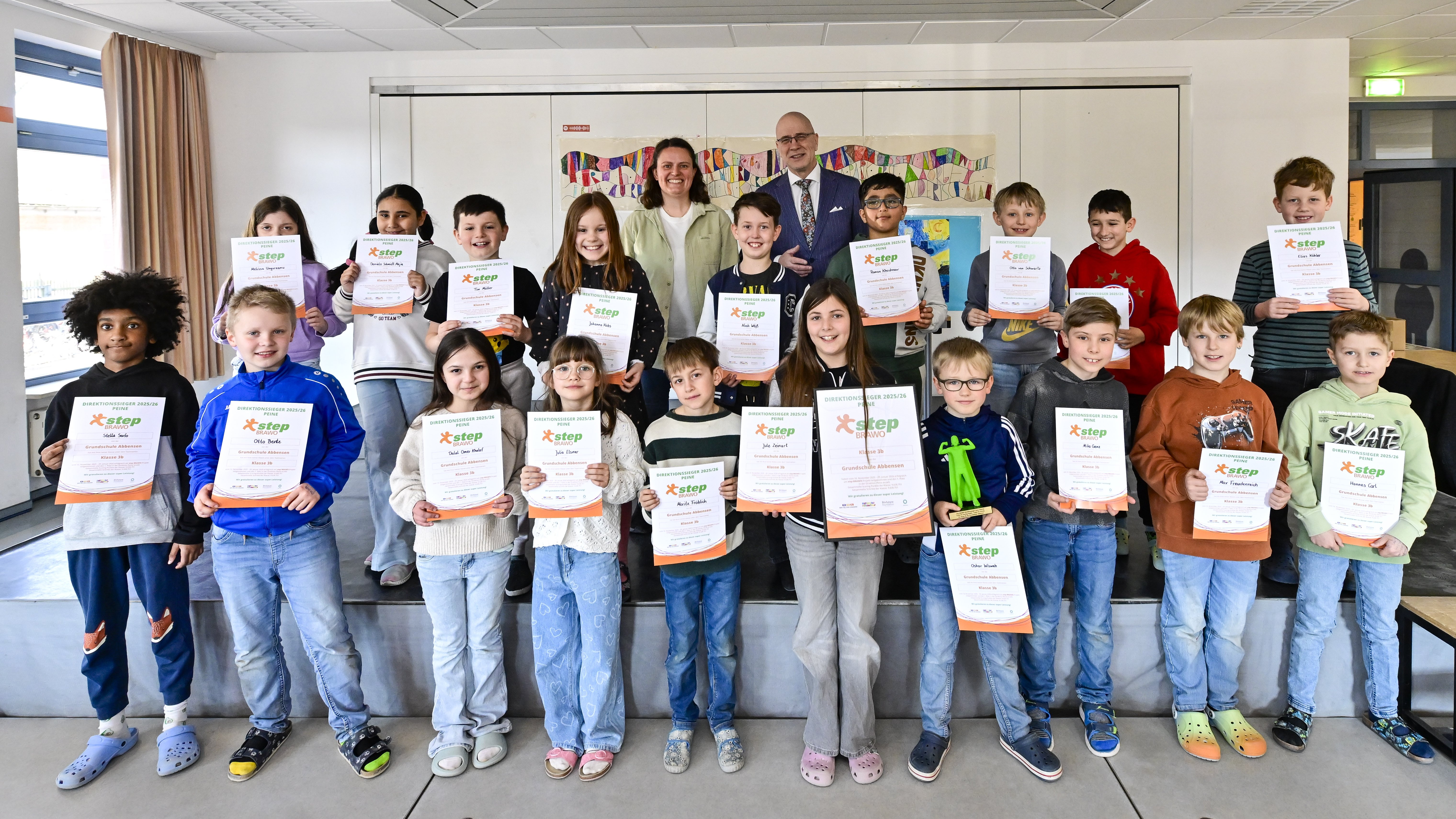 Eine Gruppe von lächelnden Kindern und zwei Erwachsenen steht in einem hellen Raum. Die Kinder halten bunte Zertifikate in den Händen. Alle wirken stolz und fröhlich.