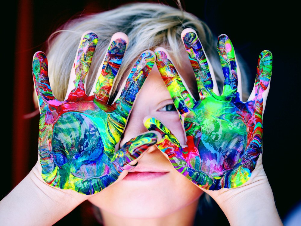 Kind hält seine Hände offen vor sein Gesicht, sodass seine bunt bemalten Handflächen zu sehen sind.