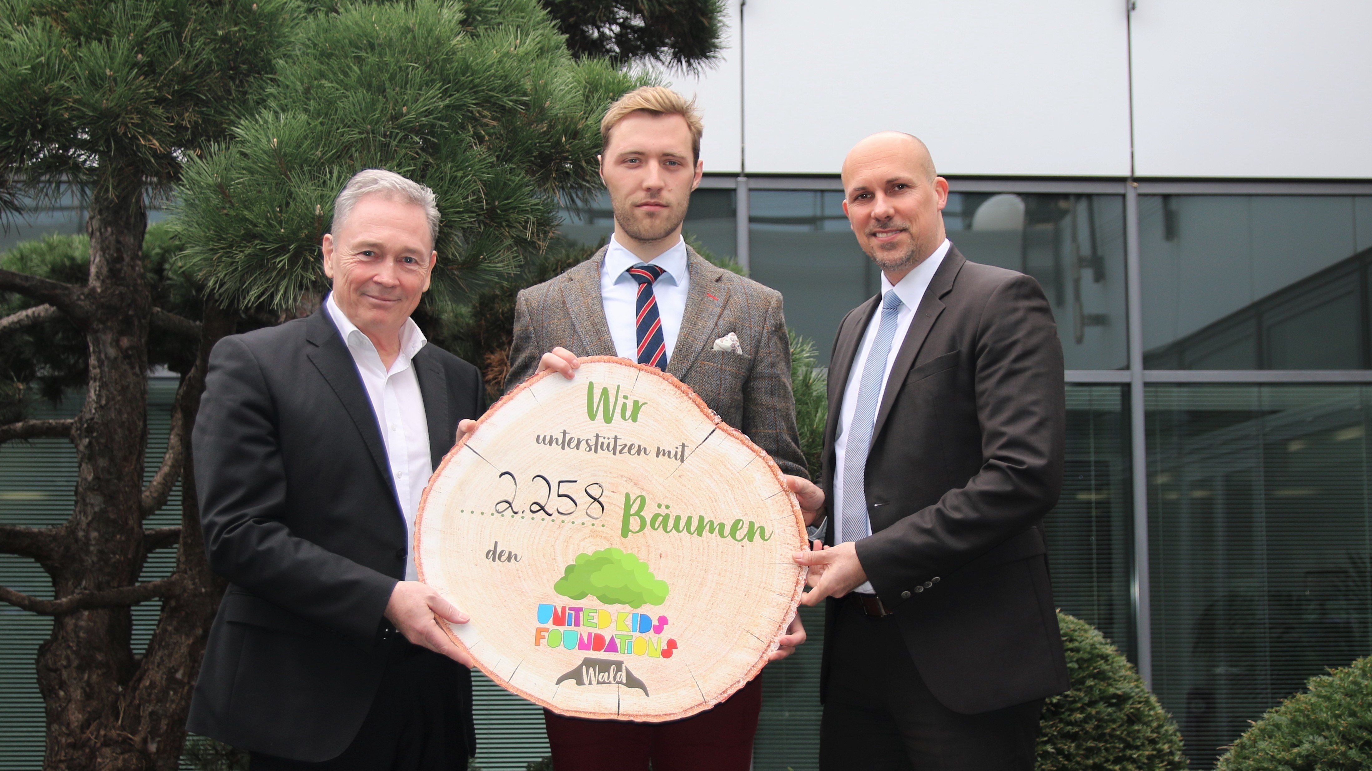 André Bonitzke (rechts), übergab im Beisein von Thomas Fast (links), ymbolisch die 2.258 Bäume für den United Kids Foundations Wald an Dr. Janis Meyerhof (mitte) von den Niedersächsischen Landesforsten. Drei Personen in Anzug stehen vor einem Gebäude und einem Baum. Sie halten ein Aufschnitt eines Baumstammes in der Hand auf dem geschrieben steht "Wir unterstützen mit 2.258 Bäumen den United Kids Foundations Wald".