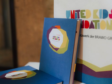 Ein Buch mit blauem Einband und buntem Logo liegt auf einem weißen Tisch. Im Hintergrund ist ein Plakat mit dem Schriftzug "United Kids Foundations" sichtbar. Das Logo auf dem Buch ist bunt und das Plakat hat einen ähnlichen farbenfrohen Stil.