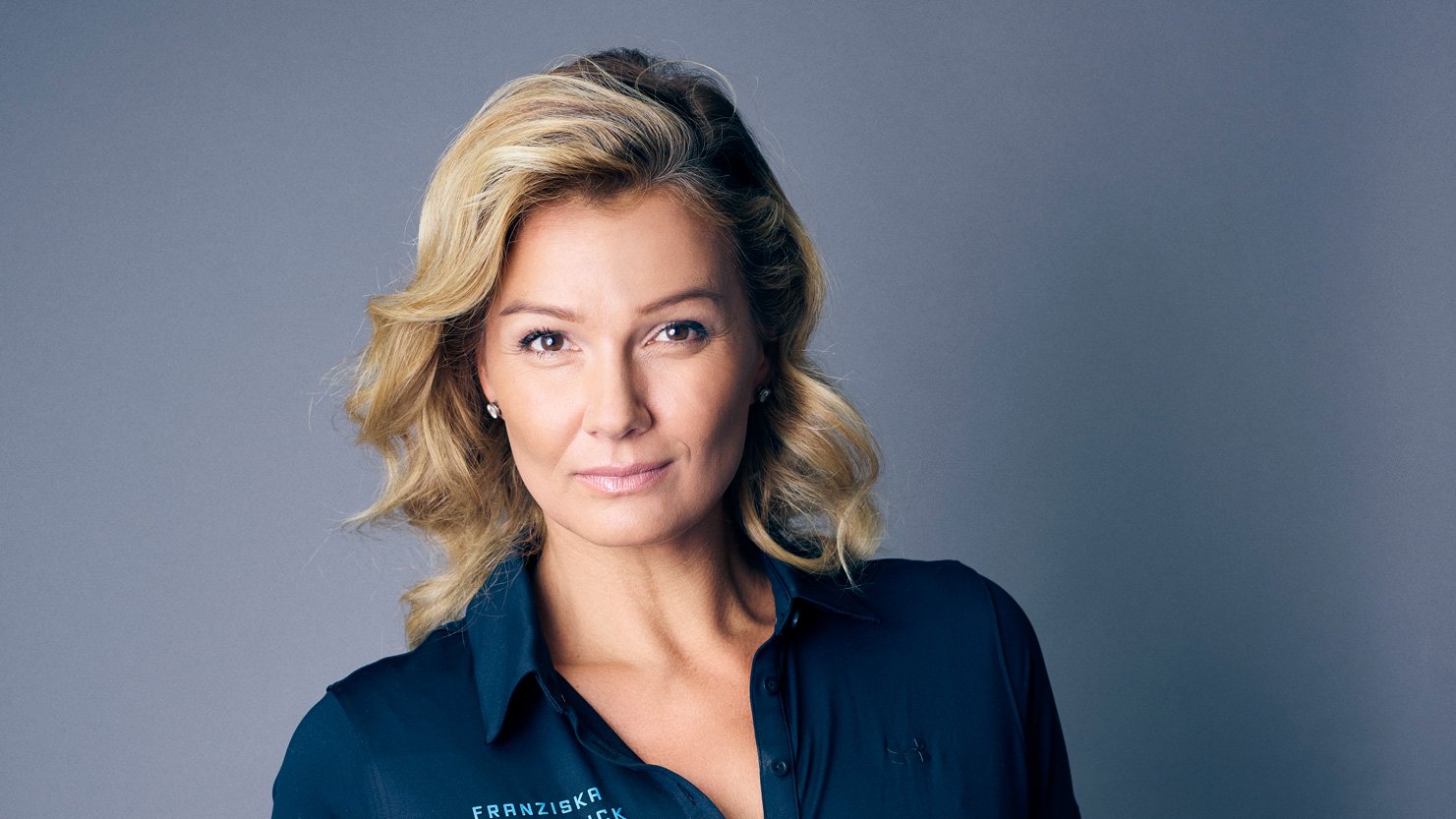 Eine Frau mit blonden, welligen Haaren steht vor einem grauen Hintergrund. Sie trägt ein dunkelblaues, kurzärmliges Polohemd mit einem Aufdruck auf der linken Seite. Ihre rechte Hand ist lässig in die Hosentasche einer blauen Jeans gesteckt. Sie hat ein warmes Lächeln und trägt ein dezentes Armband am linken Handgelenk.