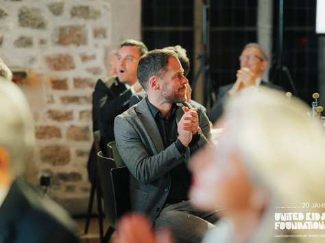 Eine Gruppe von Personen sitzt an einem Tisch in einem Veranstaltungsraum mit Steinwand. Ein Mann im Vordergrund trägt einen grauen Blazer und klatscht in die Hände. Weitere Personen im Hintergrund klatschen ebenfalls und scheinen aufmerksam zu sein. Auf dem Bild ist das Logo von "United Kids Foundations" mit dem Hinweis auf das 20-jährige Jubiläum zu sehen.