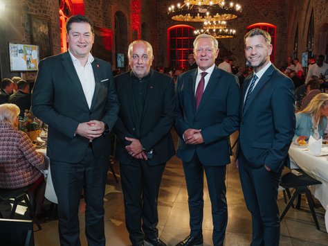 Vier Männer in Anzügen stehen in einem festlich beleuchteten Saal mit Steinwänden und großen Kronleuchtern. Sie lächeln in die Kamera. Im Hintergrund sind Menschen an Tischen zu sehen, die an einer Veranstaltung teilnehmen. Auf dem Bild ist das Logo „United Kids Foundations – 20 Jahre“ zu erkennen.