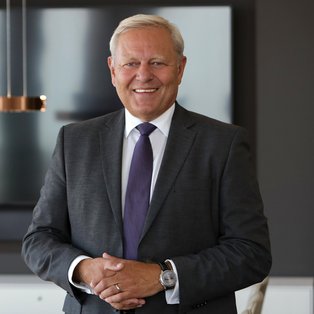 Ein lächelnder Mann in einem grauen Anzug und lila Krawatte steht in einem modern eingerichteten Raum. Im Hintergrund sind eine stylische, hängende Lampe und ein großer Bildschirm zu sehen. Der Raum wirkt hell und modern, mit weißen Möbeln.