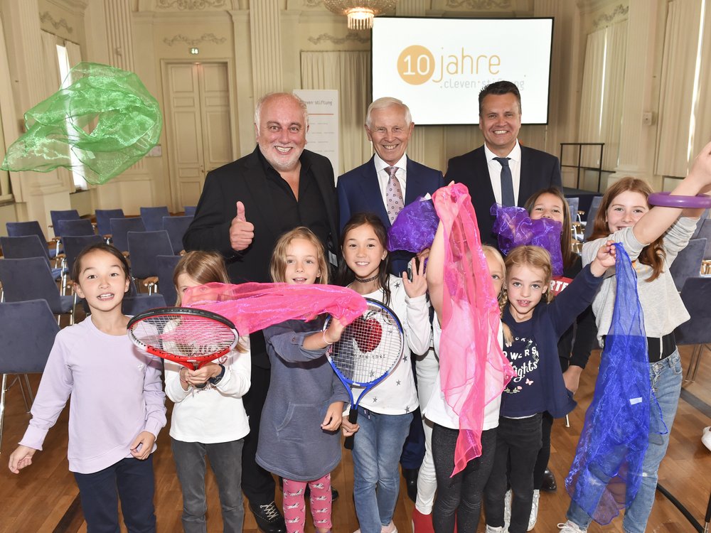 Eine Gruppe fröhlicher Kinder mit bunten Tüchern und Sportgeräten posiert mit drei Erwachsenen in einem Konferenzraum.
