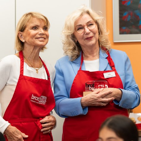Zwei Frauen stehen lächelnd nebeneinander in einer Küche. Beide tragen rote Schürzen mit der Aufschrift "brotZeit". Die Frau links hat kurzes blondes Haar und trägt eine weiße Bluse. Die Frau rechts hat mittellanges blondes Haar, eine Brille und trägt einen blauen Blazer. Im Hintergrund sind Speisen auf einem Tisch zu erkennen.