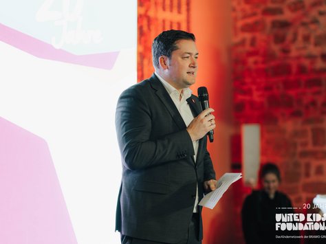 Ein Mann in Anzug und weißem Hemd hält ein Mikrofon und spricht, während er ein Blatt Papier in der anderen Hand hält. Im Hintergrund ist eine Person unscharf zu sehen. Eine große, helle Leinwand zeigt den Text "20 Jahre" und das Logo von United Kids Foundations. Die Ziegelwand im Hintergrund ist in einem warmen Rotlicht beleuchtet.