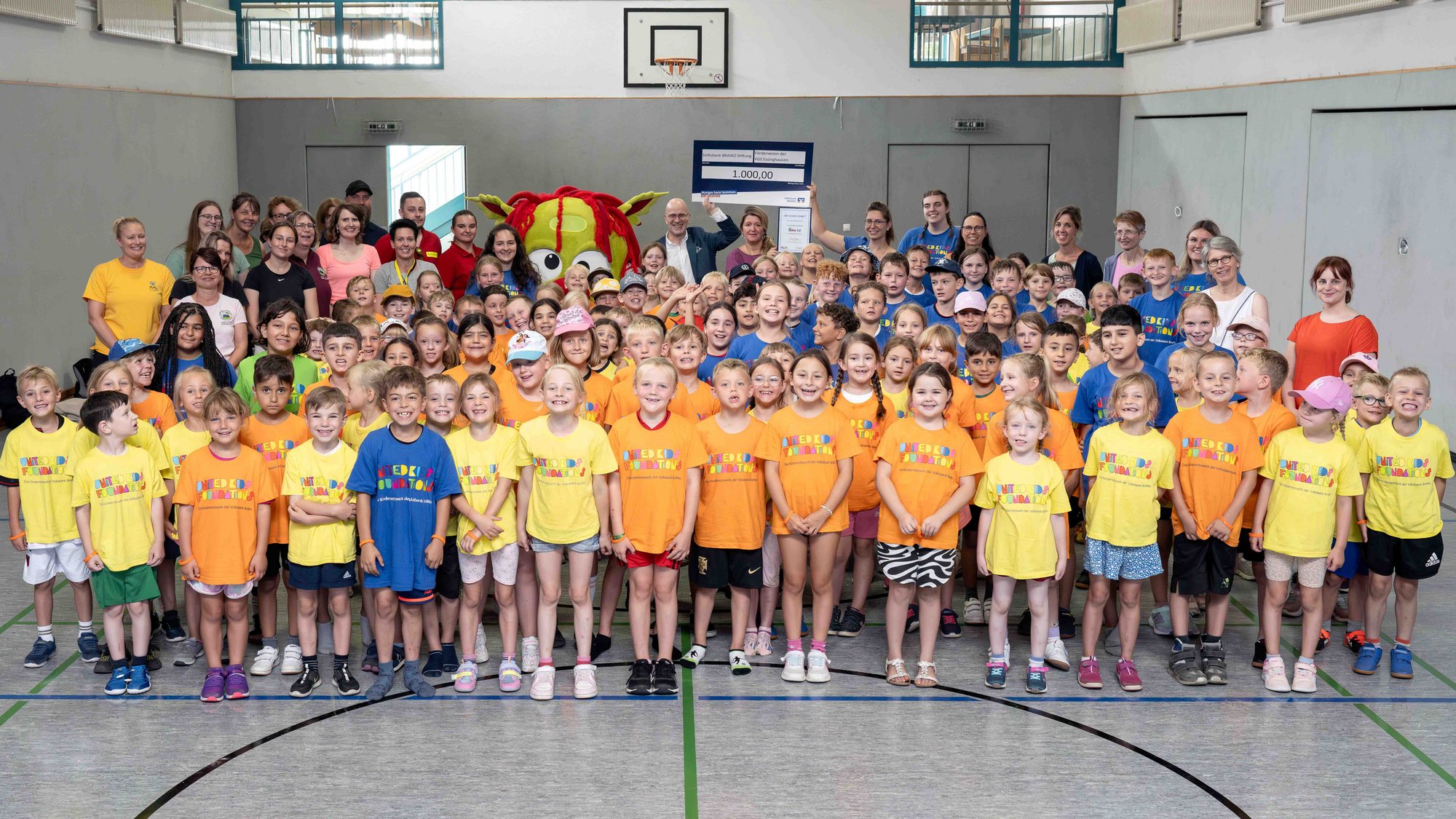 Auf diesem Bild ist eine große Gruppe Kinder, sowie vereinzelt Erwachsene in der hintersten Reihe. Die Gruppe steht in einer Turnhalle.